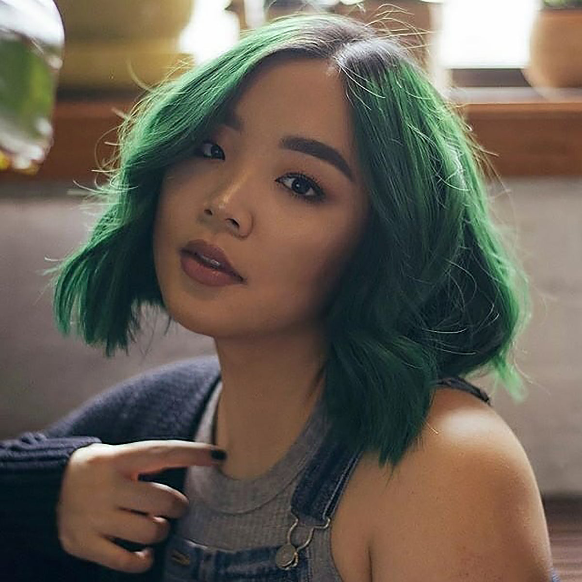 person with short, curled emerald green hair on pre-lightened hair with dark roots wearing overalls