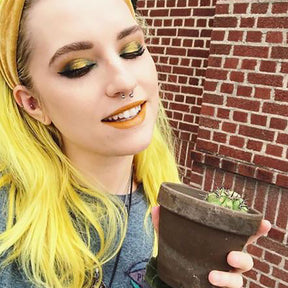 person with yellow headband and bright yellow, medium length pre-lightened hair holding a small potted cactus